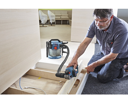 Artisan travaillant avec un rabot électrique et un aspirateur dans une pièce.
