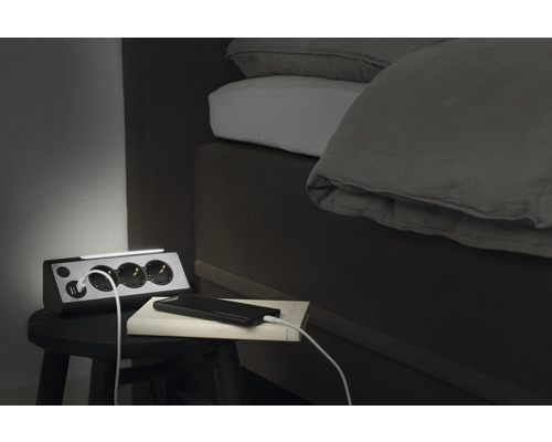 Mehrfachsteckdose mit USB-Anschlüssen und einer Lichtleiste auf einem Nachttisch neben einem Bett.