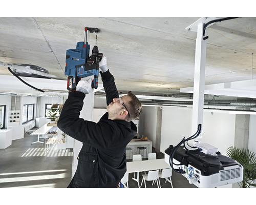 Un homme perce un trou dans le plafond avec une perceuse à percussion dans une pièce lumineuse.