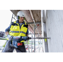 Ein Bauarbeiter bohrt mit einem Bohrhammer ein Loch in eine Betonwand auf einem Baugerüst.