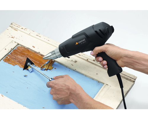 Une personne enlève de la peinture d''un cadre de fenêtre avec un pistolet thermique et un grattoir à peinture