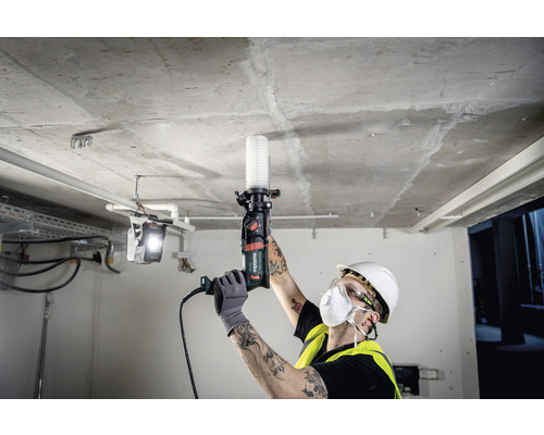 Un homme avec un casque de protection et un masque respiratoire perce un plafond avec une perceuse Metabo.