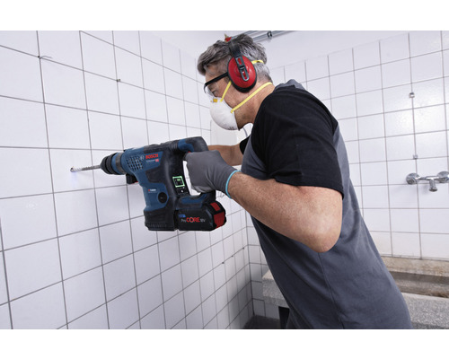Mann bohrt mit einem Bohrhammer ein Loch in eine geflieste Wand. Er trägt Schutzkleidung.