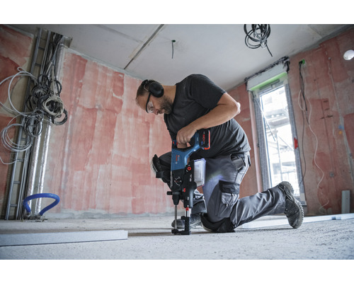 Un homme est agenouillé et perce un sol en béton avec un perforateur dans une pièce en construction brute.
