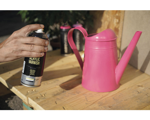 Eine Person hält eine Dose Acryllack neben einer Giesskanne auf einer Holzpalette.