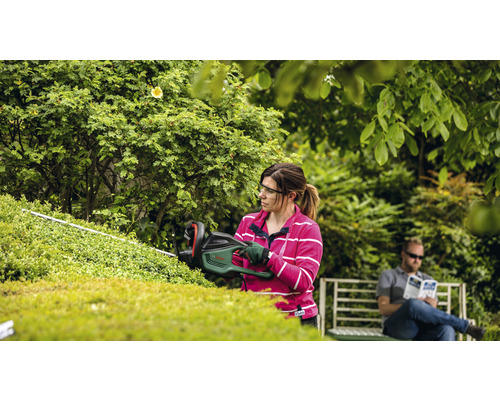 Une femme taille une haie avec un taille-haie dans le jardin, tandis qu''un homme est assis sur un banc