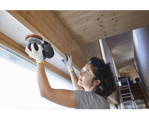 Une femme ponce un cadre de fenêtre en bois avec une ponceuse excentrique.