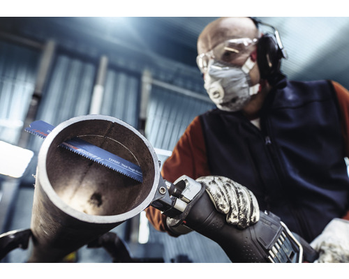 Un homme portant des lunettes de protection, un masque respiratoire, une protection auditive et des gants coupe un tuyau avec une scie sabre.