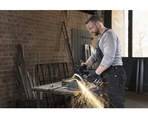 Un homme portant des lunettes de protection travaille un tube métallique avec une meuleuse d''angle dans un atelier.