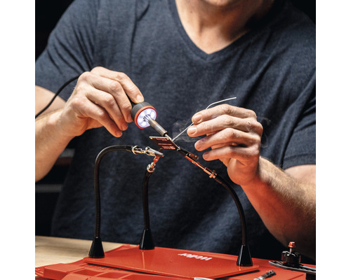 Personne en train de souder avec un outil d''aide à la soudure et un fer à souder sur une surface de travail rouge