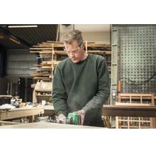 Ein Mann arbeitet mit einer Oberfräse an einem Holzstück in einer Werkstatt, er trägt dabei eine Schutzbrille.