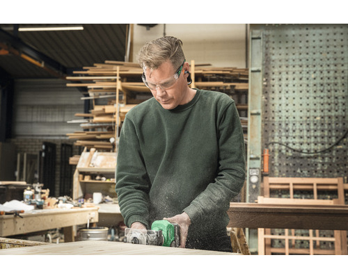 Ein Mann arbeitet mit einer Oberfräse an einem Holzstück in einer Werkstatt, er trägt dabei eine Schutzbrille.