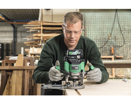 Ein Mann benutzt eine Akku-Oberfräse zum Bearbeiten eines Holzstücks. Er trägt Schutzbrille und Handschuhe.