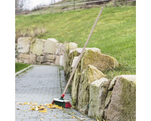 Kehrbesen mit Holzstiel lehnt an einer Steinmauer auf einem gepflasterten Weg mit Blättern