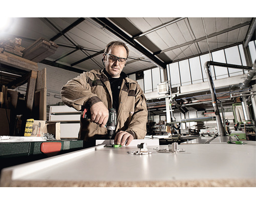 Ein Handwerker befestigt mit einem Akkuschrauber ein Beschlagteil an einer Platte in einer Werkstatt.