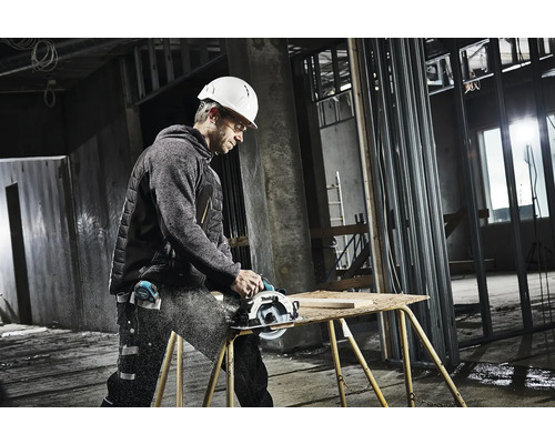 Ein Handwerker mit Schutzhelm und Schutzbrille sägt ein Holzbrett mit einer Handkreissäge auf einer Werkbank.