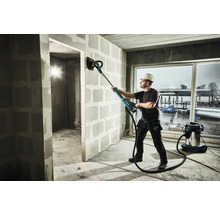 Un homme ponce un mur avec une ponceuse à long manche dans un bâtiment en construction, un aspirateur se trouve à côté.