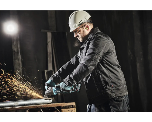 Ein Mann mit Schutzhelm, Schutzbrille und Gehörschutz schleift ein Metallrohr mit einem Winkelschleifer.
