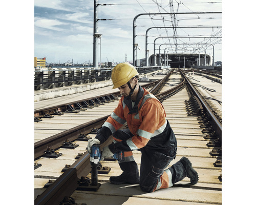 Ein Bauarbeiter mit Schutzhelm benutzt einen Akku-Schlagschrauber an Bahngleisen.