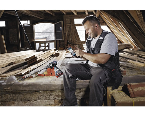Ein Mann benutzt eine Bohrmaschine und ein Smartphone auf einem Dachboden mit Holzbalken