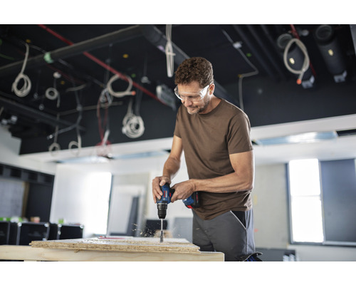 Un homme perce une planche de bois avec une perceuse. Il porte des lunettes de protection.