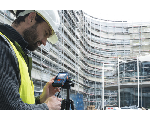 Un homme portant un casque de protection et un gilet de sécurité utilise un télémètre laser sur un trépied devant un bâtiment en construction.