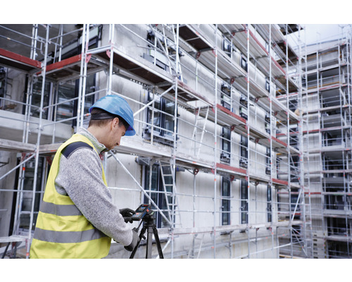 Un ouvrier du bâtiment portant un casque et un gilet de sécurité utilise un télémètre laser sur un trépied devant un bâtiment avec un échafaudage.