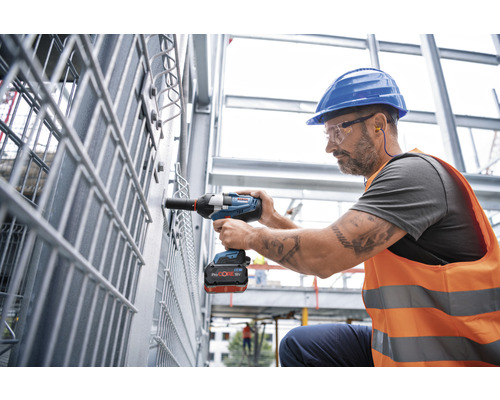 Ein Arbeiter mit Schutzhelm und Sicherheitsbrille benutzt einen Akku-Schlagschrauber an einer Metallkonstruktion.