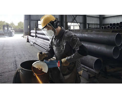 Ein Mann mit Schutzkleidung bearbeitet ein Metallrohr mit einem Winkelschleifer in einer Werkstatt.