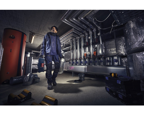 Un homme porte des outils dans un local de chauffage avec des chaudières et des tuyaux.