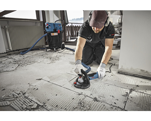 Ein Mann verwendet einen Betonschleifer auf einem Boden, der mit einem Staubsauger verbunden ist. Bosch Logo.