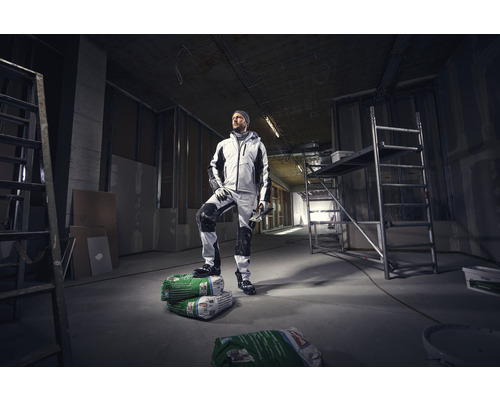 Un artisan en vêtements de travail se tient dans un bâtiment brut sur des sacs de mortier d''enduit.