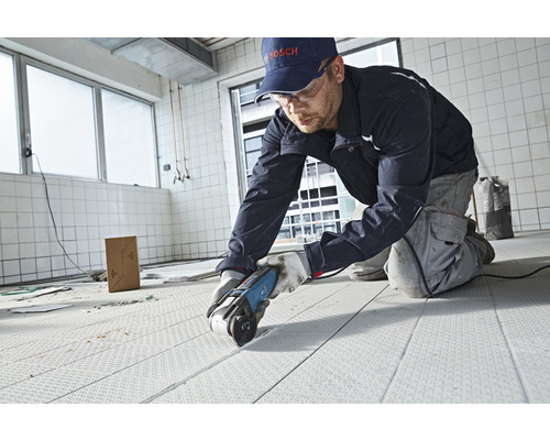Handwerker schneidet Bodenbelag mit einem Elektrowerkzeug in einem Raum. Im Hintergrund sind Fliesen zu sehen.