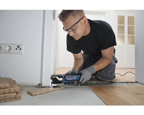 Un artisan utilise un outil multifonction pour couper un morceau de bois afin de l''adapter à une porte. L''outil est bleu et noir.