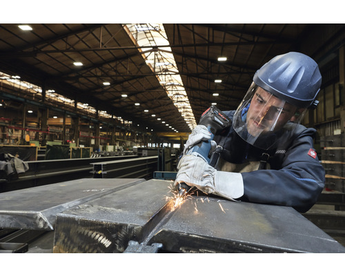 Mann schleift Metall mit einem Winkelschleifer und trägt dabei Schutzkleidung in einer Werkshalle.