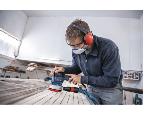 Ein Mann schleift Holz mit einem Exzenterschleifer der Marke Bosch, während er Schutzbrille und Gehörschutz trägt.