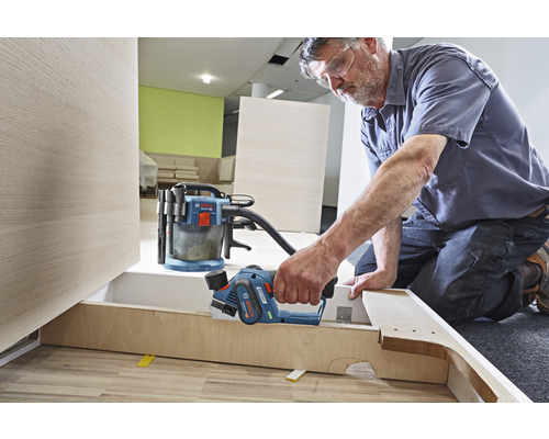 Handwerker arbeitet mit einem Hobel an einem Holzstück, ein Staubsauger steht bereit