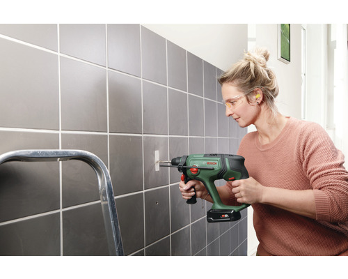 Une femme perce un trou dans un mur carrelé avec une perceuse. Elle porte des lunettes de protection et des bouchons d'oreille.