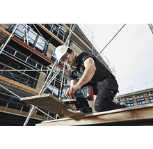 Un artisan avec un casque de protection et des lunettes de protection est en train de scier une planche en bois avec une scie sauteuse sans fil sur un échafaudage.