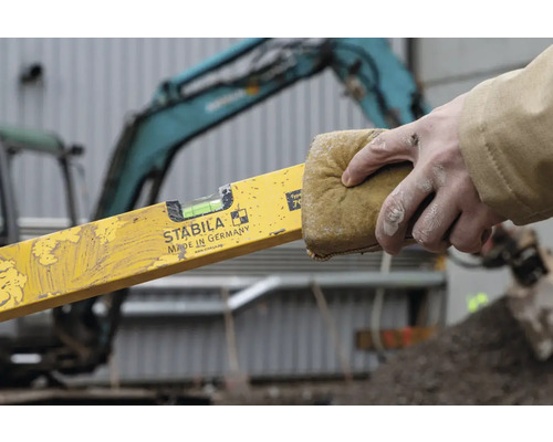 Eine Person reinigt eine gelbe Wasserwaage der Marke Stabila mit einem Schwamm. Im Hintergrund befindet sich ein Bagger.