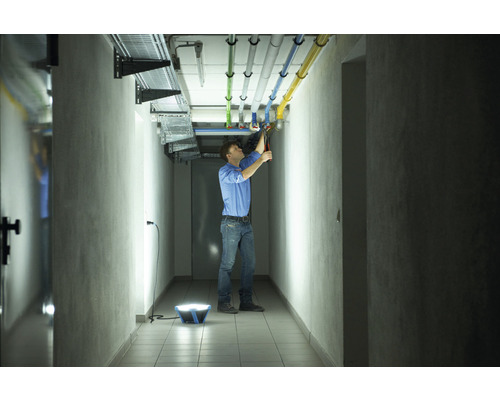 Ein Mann arbeitet in einem Flur mit einer tragbaren Arbeitsleuchte, um Rohre an der Decke zu reparieren