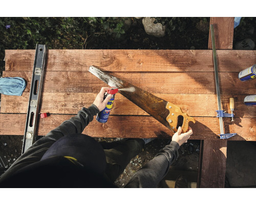 Une personne vaporise une scie à main avec un produit multifonctionnel sur une surface en bois.
