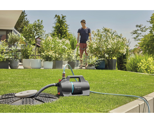 Jardin avec pelouse, sur laquelle se trouve une pompe de jardin Gardena, tandis qu''une personne arrose des plantes en arrière-plan