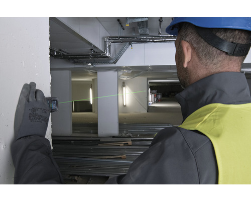 Ein Mann mit Schutzhelm und Handschuhen benutzt einen Laser-Entfernungsmesser auf einer Baustelle, um eine grüne Laserlinie auf eine Wand zu projizieren.