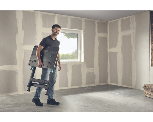 Un homme porte un établi Wolfcraft Master 650 dans une pièce avec des murs en plaques de plâtre