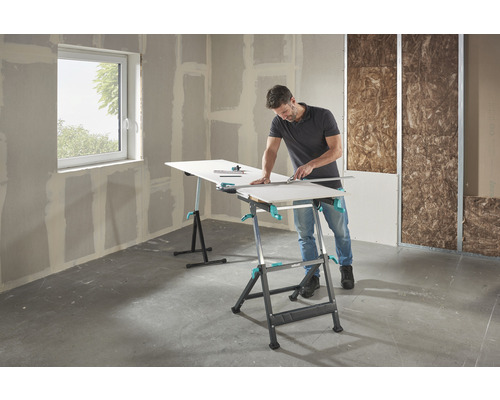 Un homme coupe une plaque de plâtre sur un établi Wolfcraft dans un bâtiment brut.