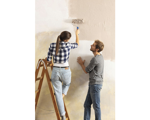 Frau streicht Wand mit Farbroller, Mann schaut zu.