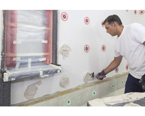 Un homme applique une isolation thermique sur un mur extérieur d''une maison, à l''aide d''une truelle et de chevilles isolantes.