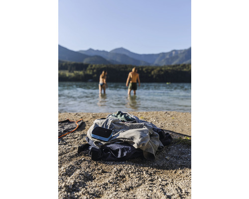 Tas de vêtements et haut-parleur sur la plage avec des personnes dans l''eau et des montagnes en arrière-plan