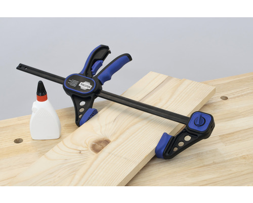 Un serre-joint à une main INDUSTRIAL fixe une planche de bois sur un établi, à côté d''une bouteille de colle à bois.